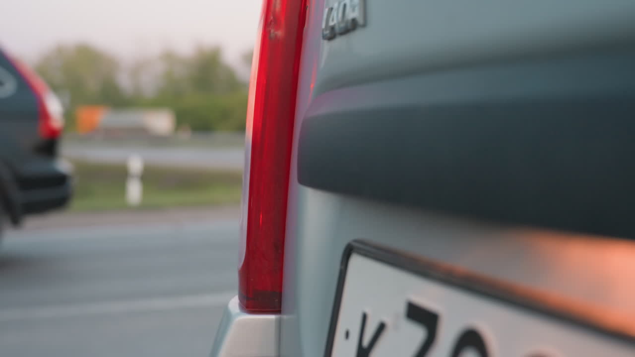 Close up rear of parked car with side light blinking near roadside, license plate partly visible, red taillight glowing under soft daylight with blurred trees and highway background