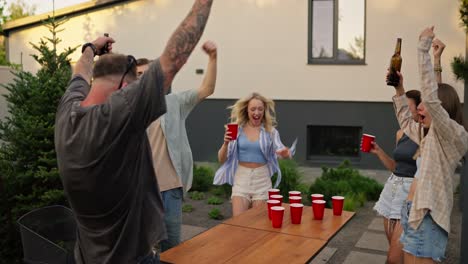 Confident-guy-in-a-gray-T-shirt-throws-a-round-white-ball-and-hits-a-cup-with-an-alcoholic-drink-while-playing-at-a-party-in-a-country-house