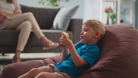 little-child-sits-in-bright-room-on-armchair,-eats-sandwich-next-to-his-mother-sitting-on-couch