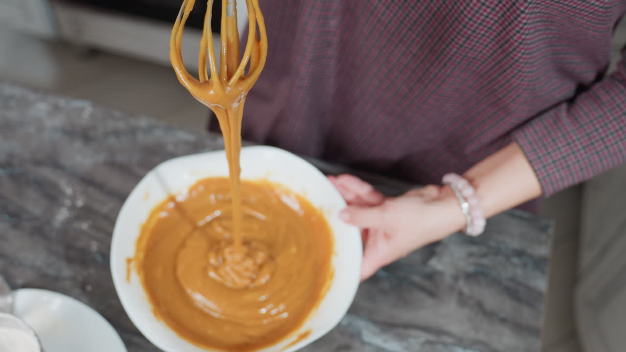 Top-down view of cook lifting whisk with thick caramel-colored pap stretching as it drips into white bowl, flour-dusted countertop and metal sifter nearby