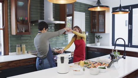 Feliz-Pareja-De-Lesbianas-Caucásicas-Preparando-Comida,-Bailando-Y-Usando-Una-Tableta-En-Una-Cocina-Soleada