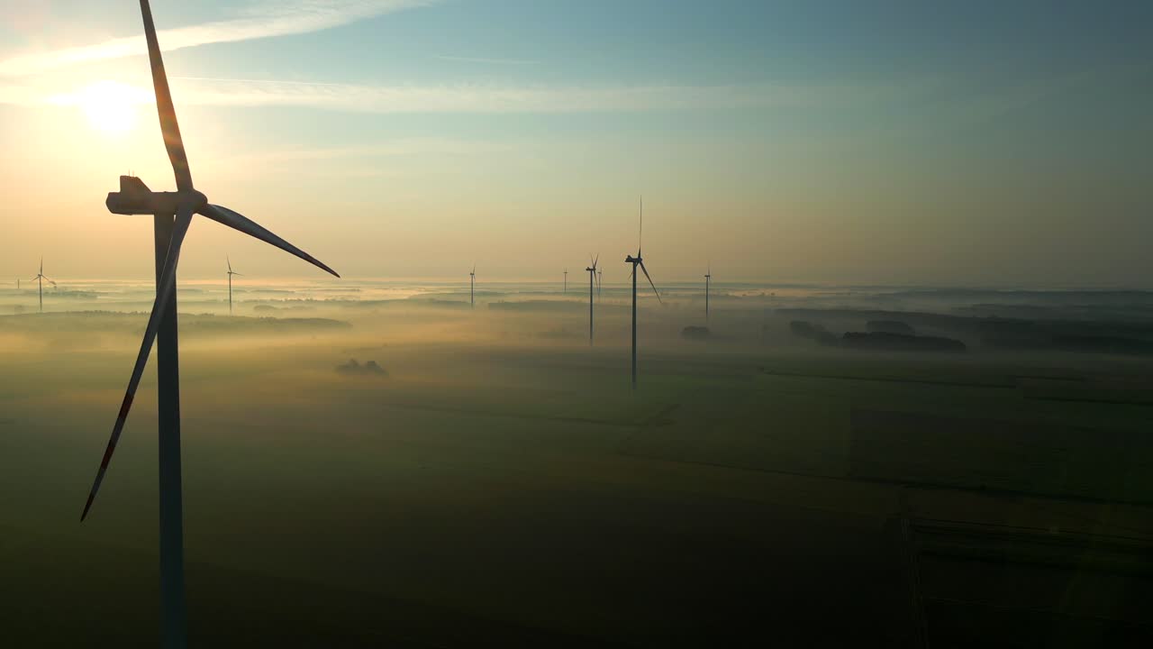 Drone's camera flies past a majestically rotating wind turbine over a landscape immersed in fog and illuminated by the setting sun