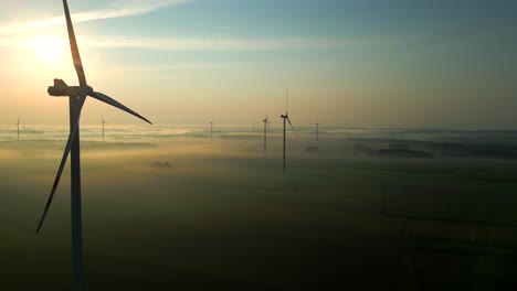 Drone's-camera-flies-past-a-majestically-rotating-wind-turbine-over-a-landscape-immersed-in-fog-and-illuminated-by-the-setting-sun