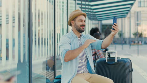 caucasian young happy man traveller sitting in bus stop talking, waving hands and smiling at the camera outdoors in summer