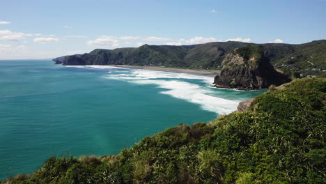 Volando-Sobre-El-Promontorio-Para-Revelar-El-Agua-Azul-Turquesa-Estrellándose-En-La-Playa-De-Piha,-Nueva-Zelanda