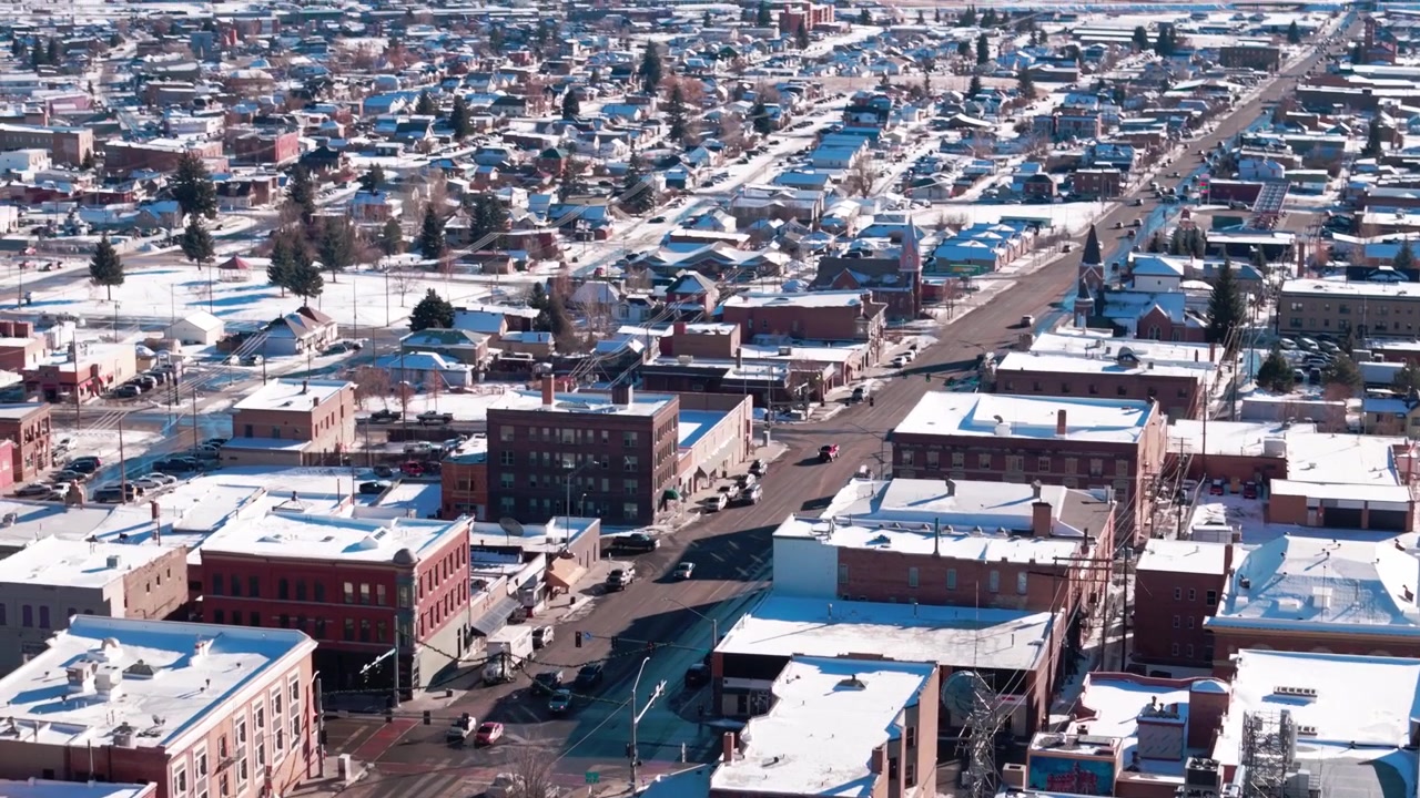 Aerial winter flight over the historic mining landscapes of Butte, Montana, showcasing closely packed snow-laden commercial buildings, vintage architecture, and surrounding snowy hills