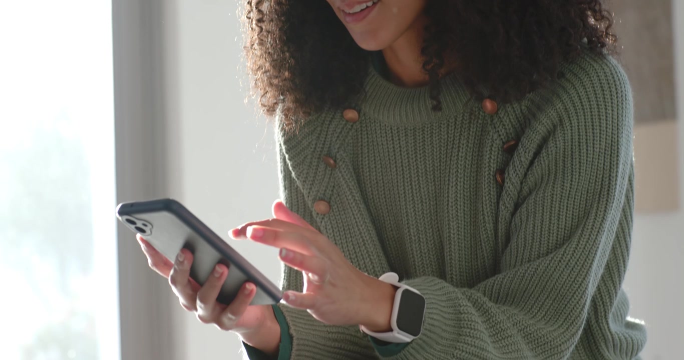 African American woman seeing alert, tapping phone, reading by window in green sweater, white watch