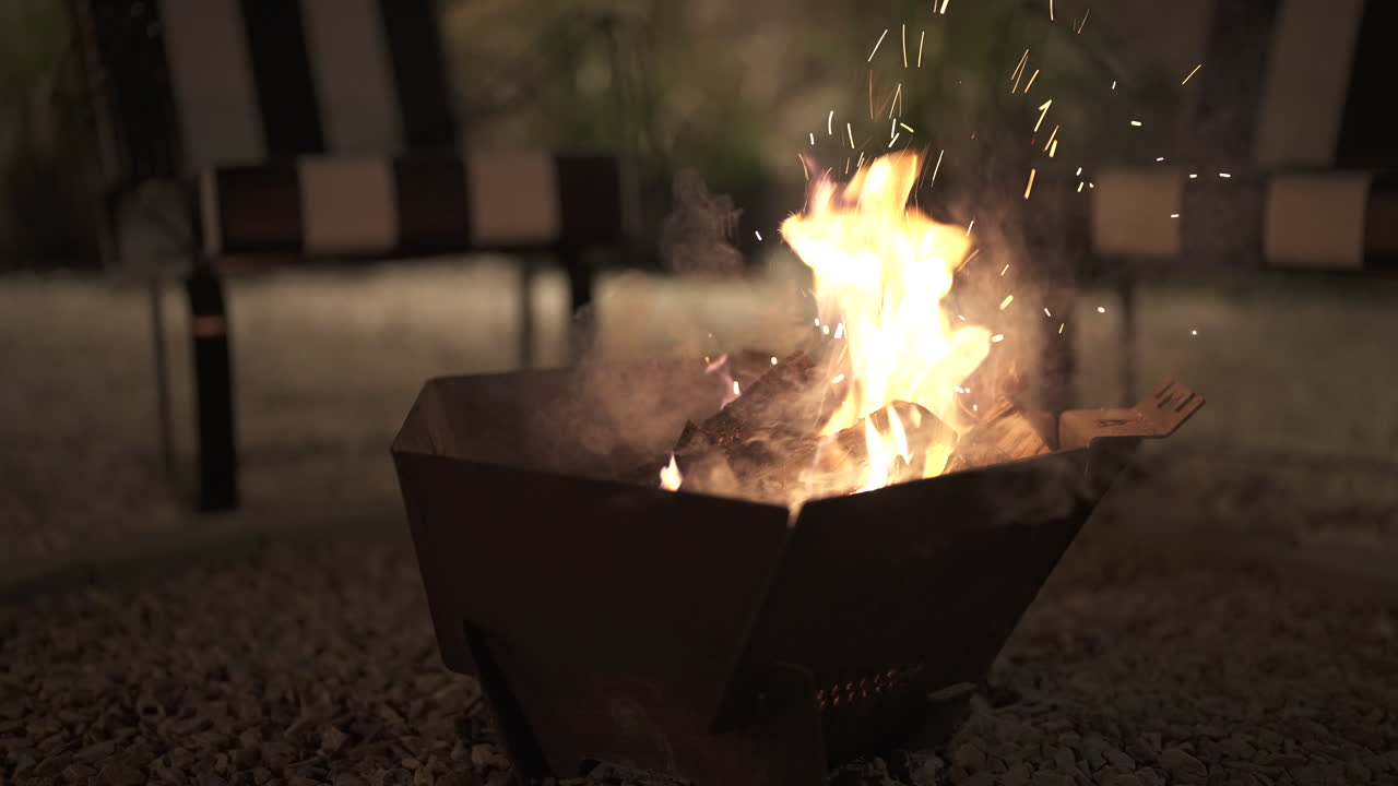 Bålpanne med brennende kubber skaper en varm og innbydende glød og sender gnister ut i den mørke natten, noe som gir en koselig atmosfære for utendørsaktiviteter på en campingplass og tilbereding av deilige marshmallows