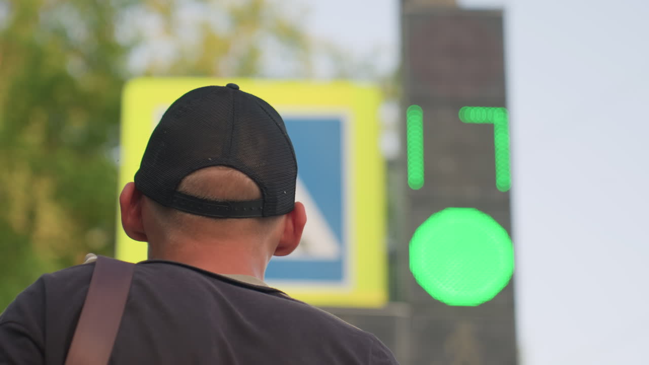 Caucasian Man Watching Traffic Signal From Rear, Cap And Backpack Strap Visible, Green Light And Red Indicators In Frame, Digital Timer, Contemplative Stance With OcdLike Fixation And Inner Conflict