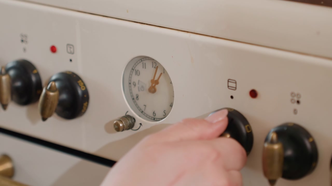 Un primo piano ravvicinato mostra una mano che afferra una manopola di controllo nera del forno e la gira su un pannello bianco del fornello per cambiare l'impostazione del calore, con una leggera sfocatura di movimento, pressione delle dita e un chiaro allineamento del quadrante.