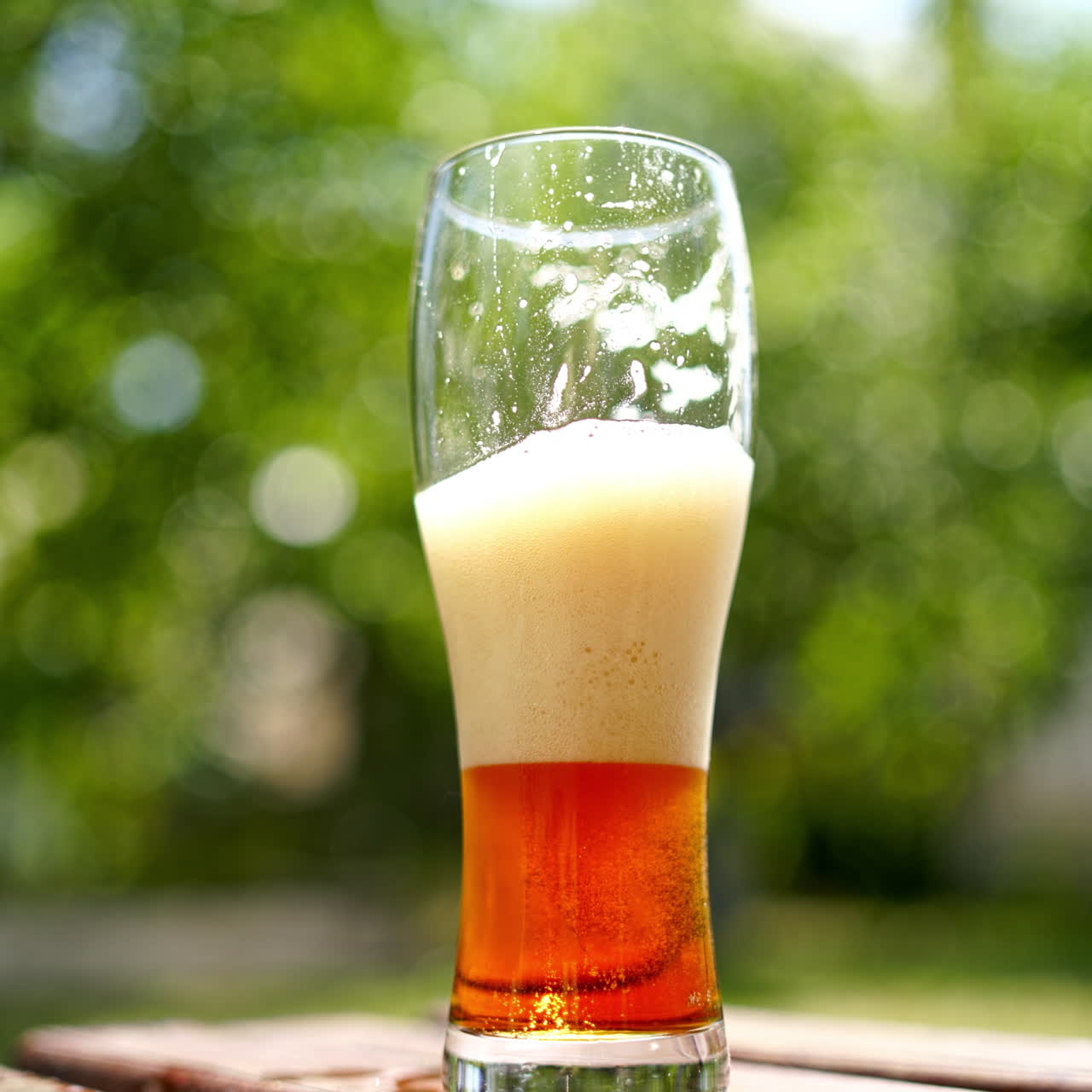 Half glass of cold beer with white foam outdoors. Close-up dark beer with bubbles on blur green background in summer.