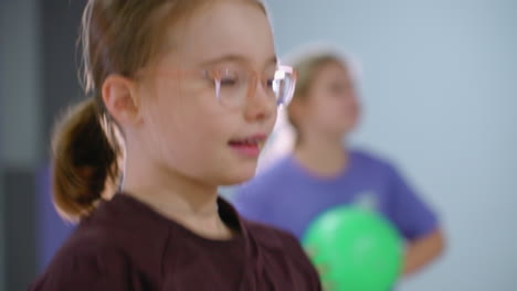 close-up of little girl in glasses juggling, throwing green balloon up, with bokeh view of another kid holding a green balloon in background, gym setting