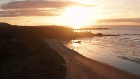 Drone-descending-over-Bastendorff-Beach-near-Coos-Bay-Oregon,-showing-Cape-Arago-lighthouse-in-the-background-as-a-silhouette