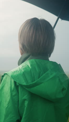 back view of woman in green raincoat holding an umbrella while standing in a vast grassy field, looking into the distance, the sky is cloudy, and trees can be seen far in the background