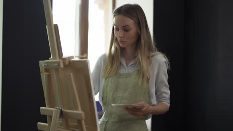 Young-woman-artist-in-apron-painting-picture-on-canvas-in-art-studio