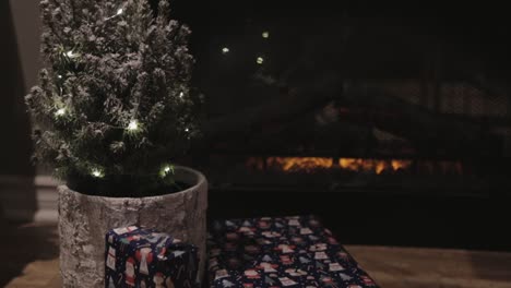 a small christmas tree with lights in a pot seated on a wooden floor near the fireplace at night- close up shot