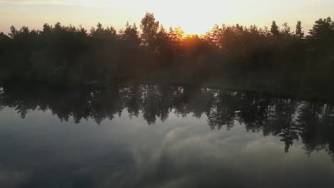 Aerial-skims-lake-surface-to-foggy-shoreline-trees-and-golden-sunrise