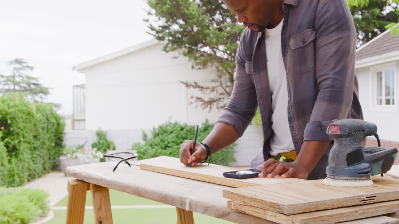 Uomo africano che posiziona una tavola sui cavalletti, con una cintura portautensili, che segna per il taglio e controlla il telefono in giardino.