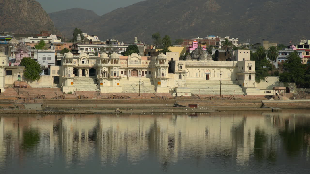 Pengambilan gambar luas Danau Pushkar dengan istana bersejarah dan ghat yang terpantul di air tenang, Rajasthan, India. Arsitektur Hindu tradisional dan tepi air suci di cahaya pagi