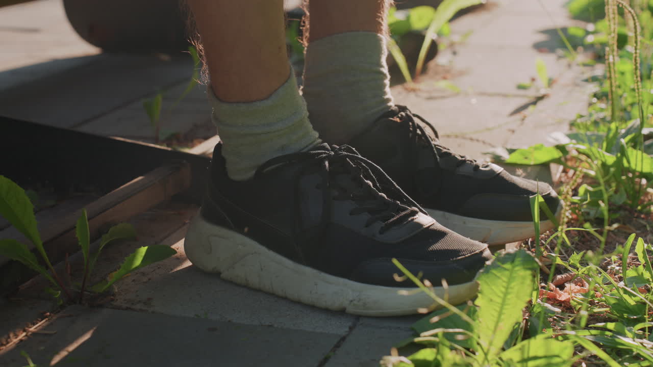 CloseUp White Feet Near Drain Grate, Black Sneakers Resting On Tiled Curb, Green Weeds And Sunlight, Quiet Stillness And Waiting, Tense Alert Posture Suggesting Paranoia And Ocd, Gritty Urban Detail