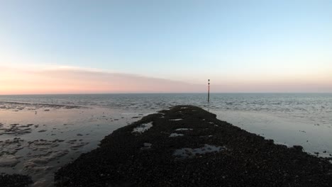 Drohnen-Luftaufnahmen-Vom-Ufer-Zum-Ozean-Mit-Bei-Sonnenaufgang-Mit-Einem-Orangefarbenen-Himmel-Mit-Einer-Stange-In-Der-Mitte