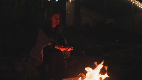 pensive young woman moves fingers over campfire. woman with blanket over shoulders strives keeping warm during chilly night in glamping house yard