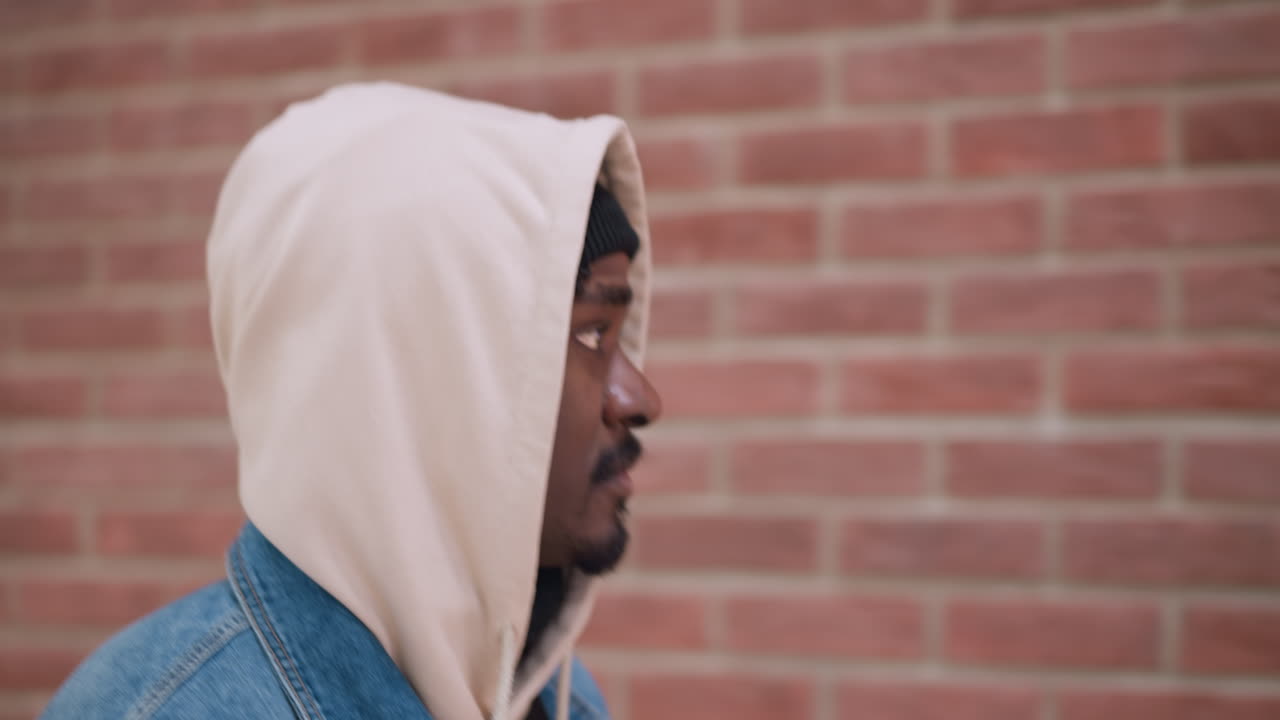 closeup portrait black man wearing beige hoodie denim jacket looking up against brick wall background expression thoughtful urban street style cinematic shot moody lighting reflection