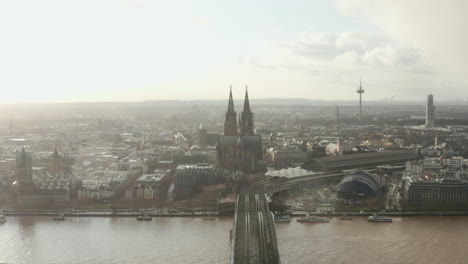 Aerial-panoramic-footage-of-town-against-sunset-sky.-Railway-bridge-over-river,-train-station-and-historic-Cologne-Cathedral.-Cologne,-Germany