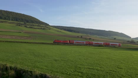 Drone-shot-at-dusk-reveals-a-red-train-with-several-passenger-cars,-moving-from-right-to-left-across-a-single-track-in-a-green-field,-a-quaint-village-with-white-houses,-under-a-pale-blue-sky