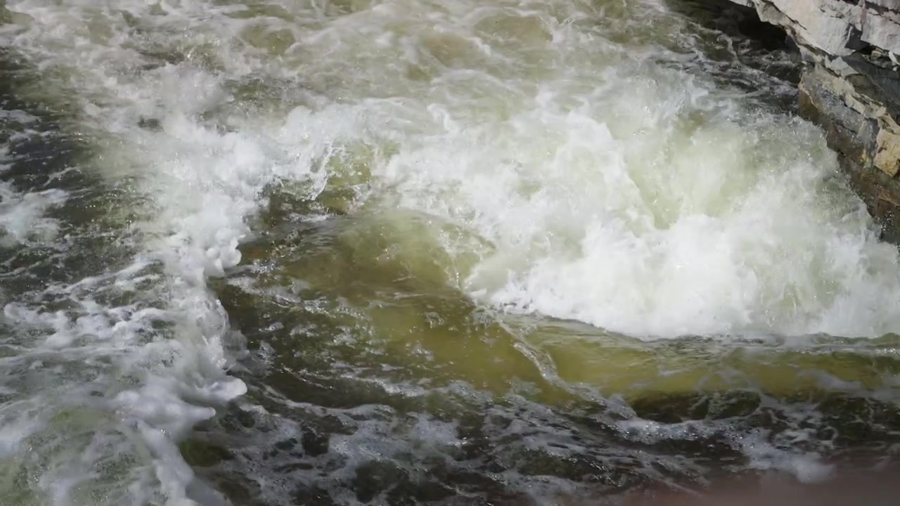 Un primo piano cattura un violento vortice di acqua bianca aerata che si agita alla base di una cascata. Le potenti correnti e la schiuma spumeggiante mostrano la pura forza della natura.