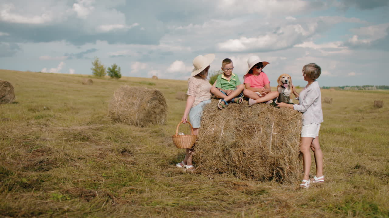 семья наслаждается пикником на открытом воздухе на стоге сена, бабушка кормит собаку, в то время как девочка протягивает руку, женщина держит корзину, дети в солнечных шляпах играют, сельский пейзаж с разбросанными балами сена