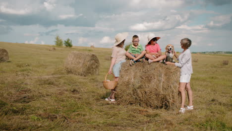 la familia disfruta de un picnic al aire libre en un pajar, la abuela alimenta al perro mientras la niña se acerca, la mujer sostiene la canasta, los niños con sombreros de sol juegan, el paisaje rural con balas de heno esparcidas