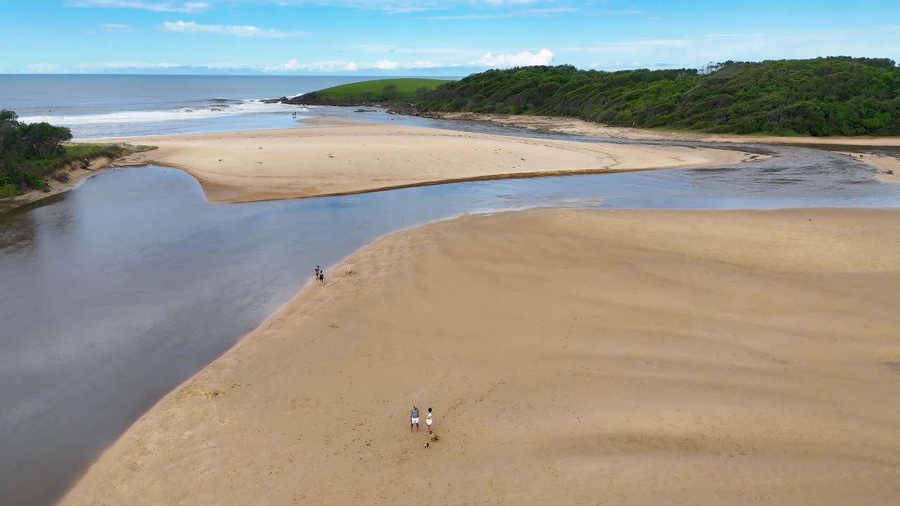 Drone tracks people walking on sandy river mouth at Moonee Beach under bright daylight
