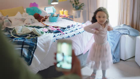 happy little girl playing dress up wearing fairy wings in bedroom with mother taking photo using smartphone sharing on social media