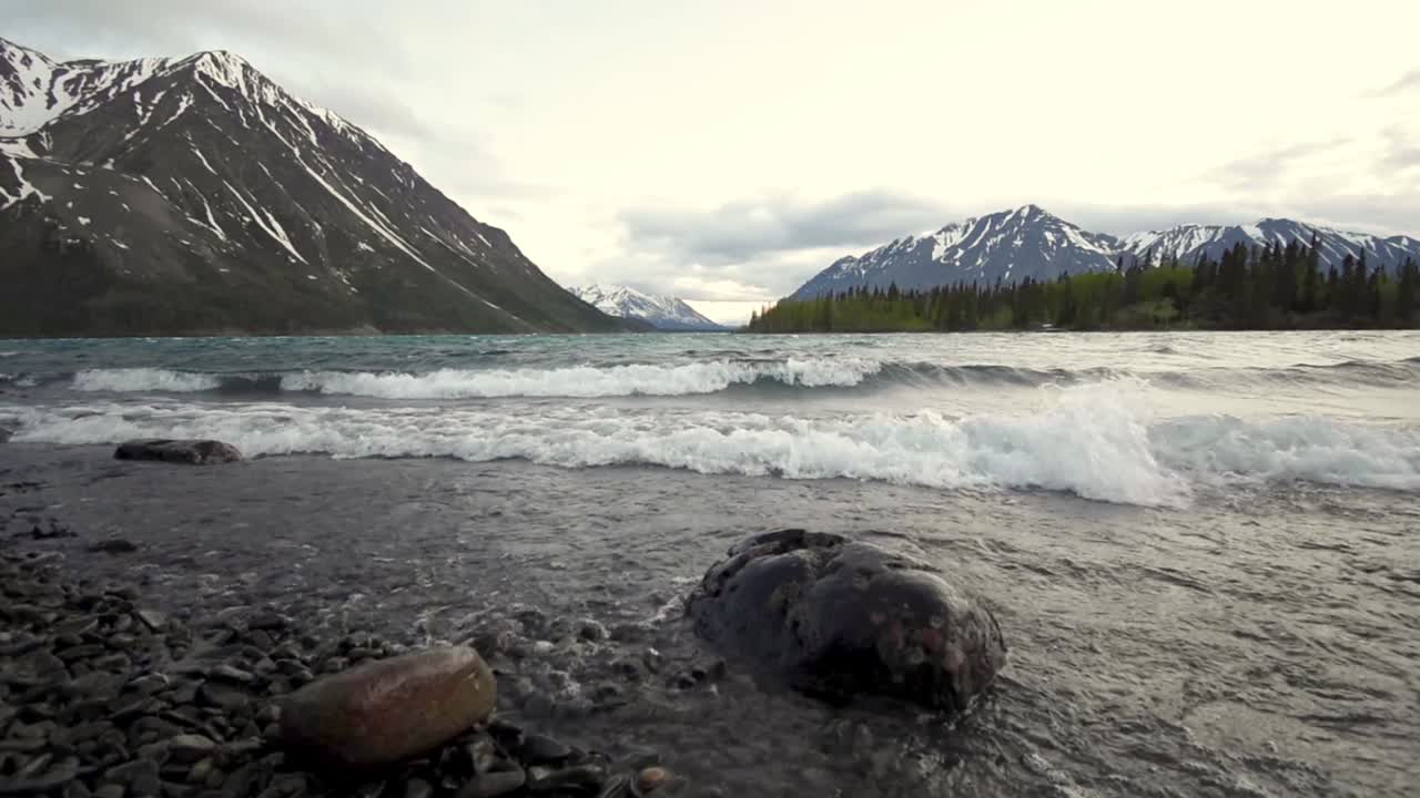 Low angle view of Yukon Kathleen lake waves rolling onto rocky shore with scenic snow capped mountains in background, Canada, wide angle static