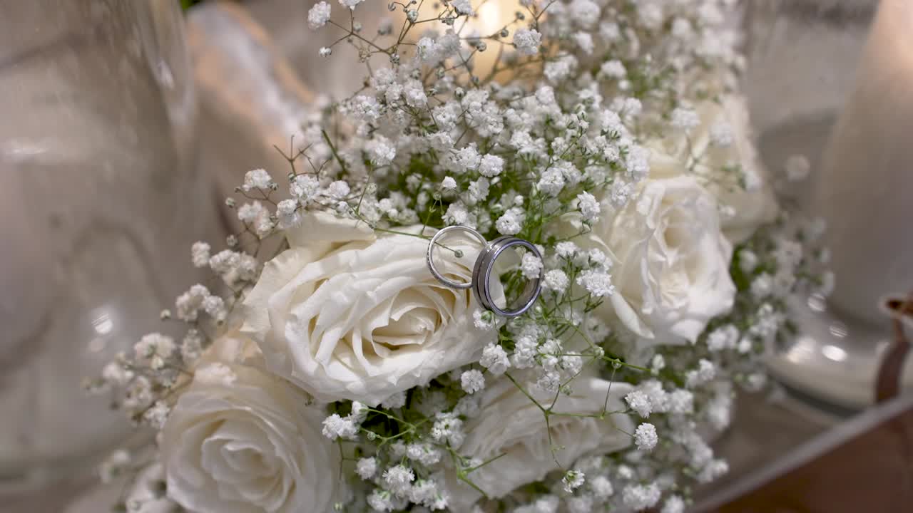 Two wedding rings rest among white roses and baby's breath in a floral arrangement. The flowers are a part of a wedding decoration set. This moment captures love and commitment