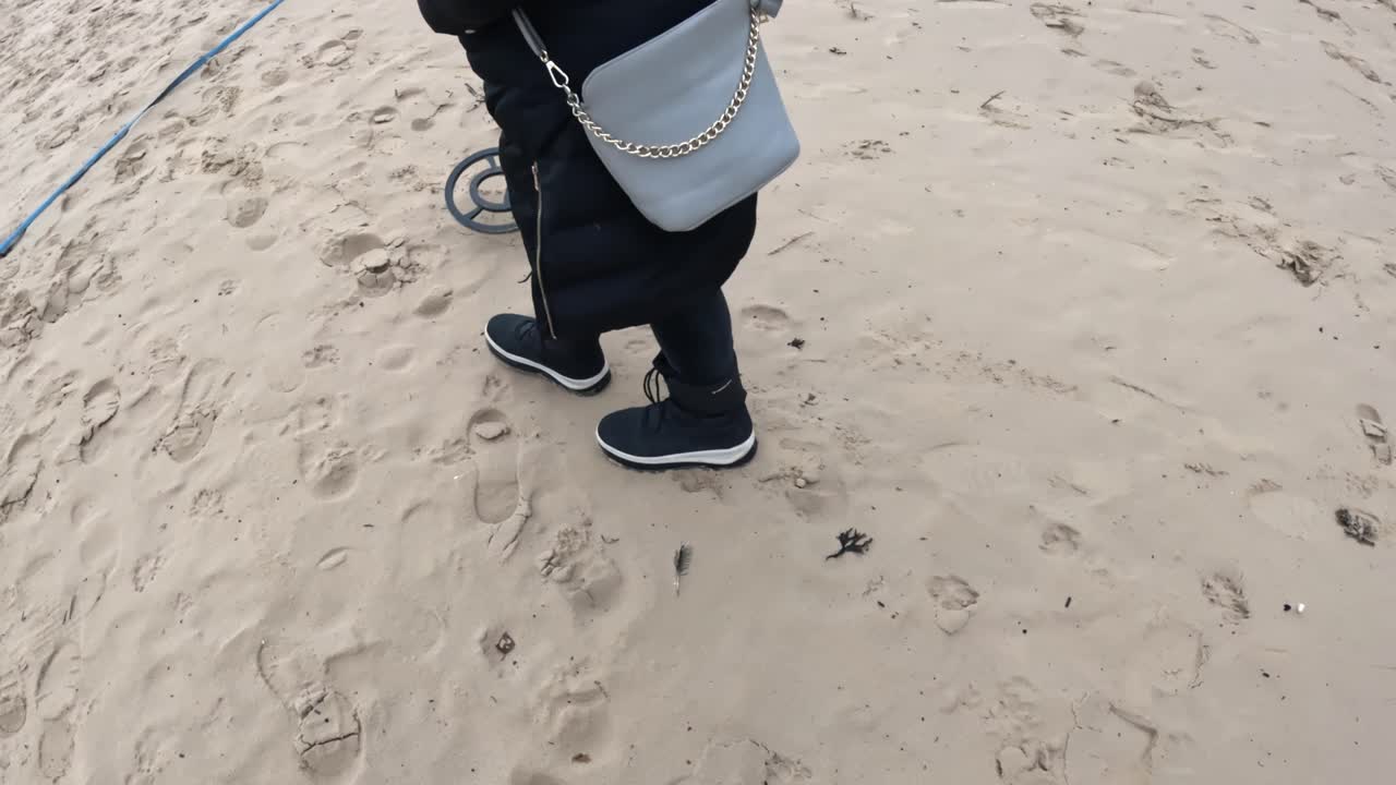 Persona che cammina sulla sabbia soffice della spiaggia mentre passa un metal detector su impronte sparse, alla ricerca di oggetti smarriti durante una tranquilla gita invernale
