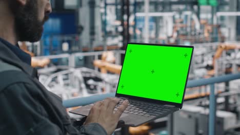 ingeniero de fábrica de automóviles en uniforme de trabajo usando una computadora portátil con pantalla verde. trabajando con software en una instalación de fabricación industrial automotriz dedicada a la producción de vehículos.