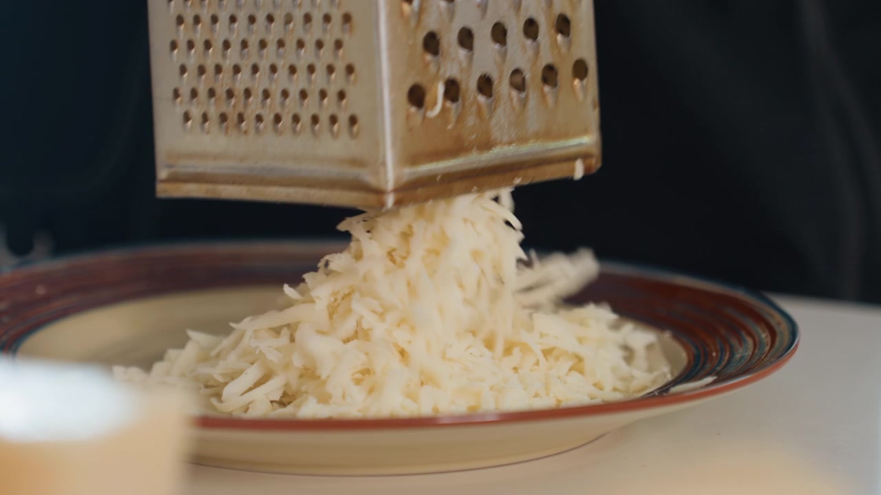 Two hands gently press and lift freshly grated white cheese on ceramic plate in kitchen, creating soft collapse, loose shreds, subtle movement, and warm indoor food preparation atmosphere with