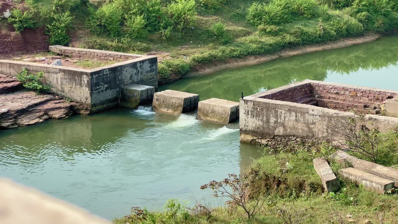 Close-up view of a small concrete dam built across a calm river in a rural area, surrounded by green vegetation and rocks, showing controlled water flow and simple irrigation infrastructure