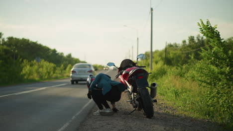 a lady wearing a helmet and checkered shirt walks along a rural road toward her parked red power bike, she rest her back on the bike, as a car passes by in the background