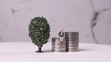 a miniature man reading a book on a pile of coins and a miniature tree.