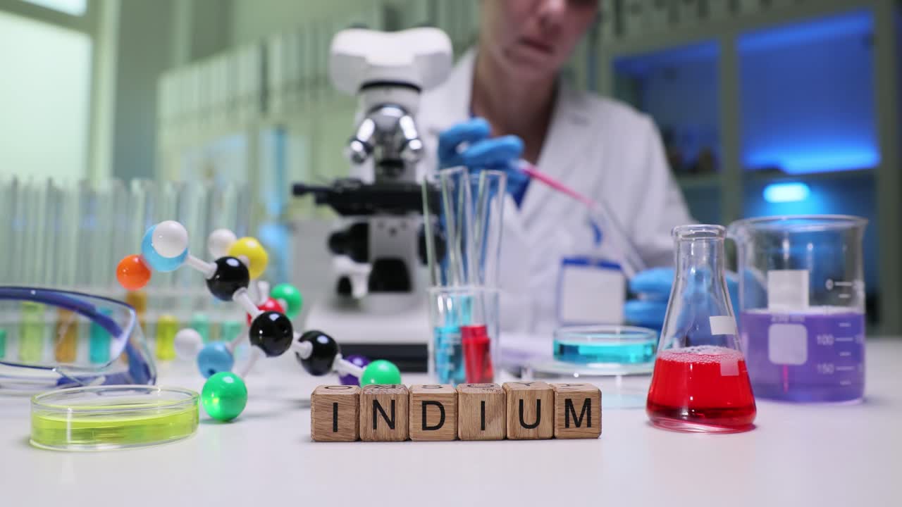 A scientist works in a lab with various chemical equipment and a visible "INDIUM" sign