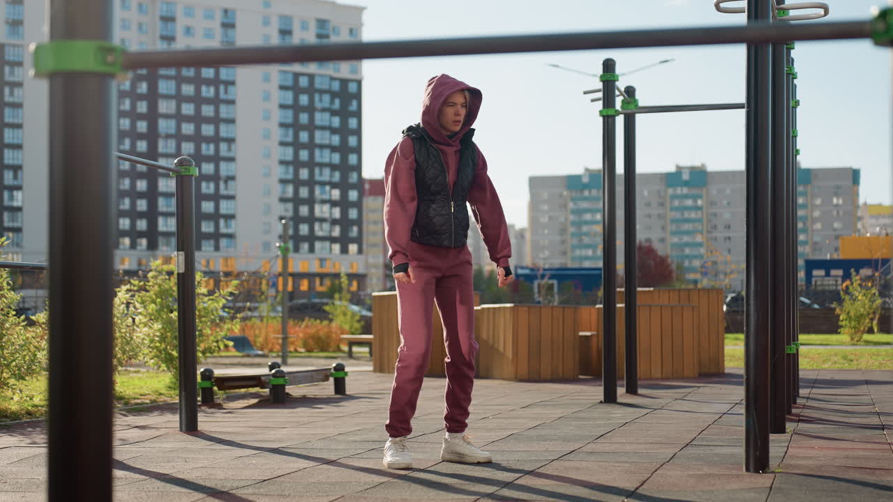 Training partner in hoodie and vest gently stretches one arm across chest with focused expression while people move in urban street with modern buildings under sun light for warm up workout session