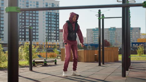 training partner in hoodie and vest gently stretches one arm across chest with focused expression while people move in urban street with modern buildings under sun light for warm up workout session