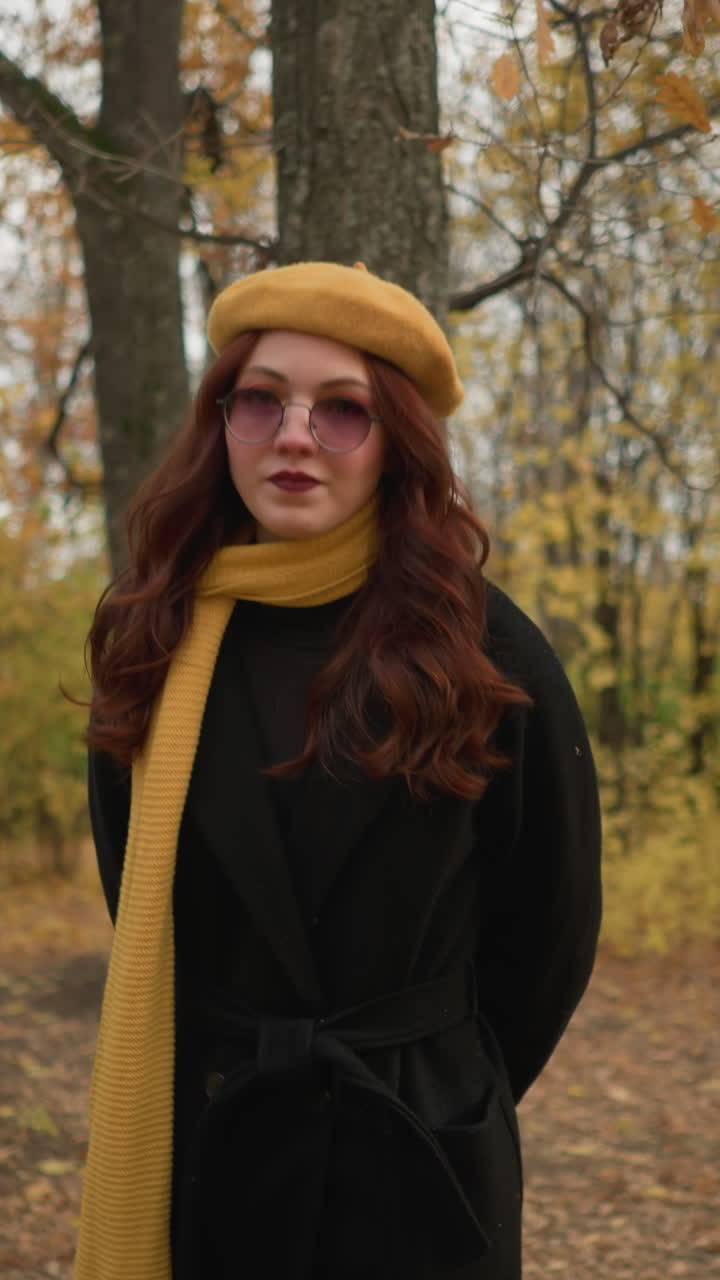 스타일리시한 노란색 베레트와 스카프를 입은 젊은 여인이 조용한 가을 숲을 걸어다니며, 바람에 부드럽게 흔들리는 잎을 관찰하고, 자연의 평화로운 분위기를 즐기고 있습니다.