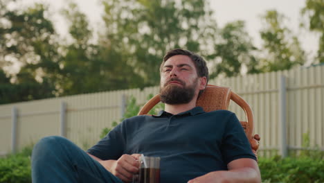 man relaxed on wicker rocker chair holding clear glass mug of hot coffee with hand close view of steam rising against blurred garden fence and bokeh tree background