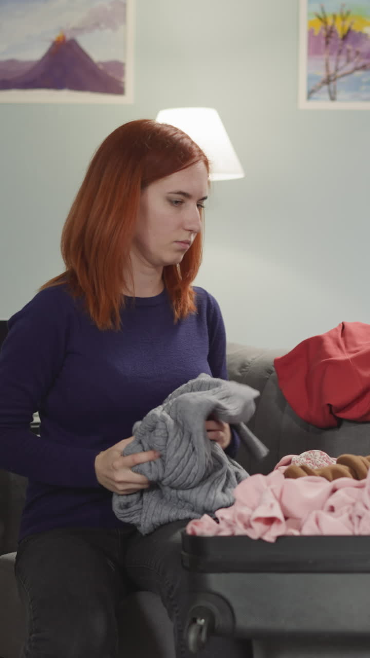 Angry redhead woman collects things after conflict with husband. Disappointed wife puts clothes into suitcase sitting on sofa in living room slow motion