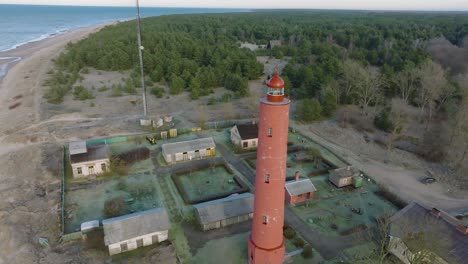 aerial establishing view of red colored akmenrags lighthouse, baltic sea coastline, latvia, white sand beach, calm sea, sunny day with clouds, wide drone orbit shot