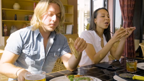 hombre rubio y mujer morena comiendo pizza sentados en una mesa de restaurante con sus amigos